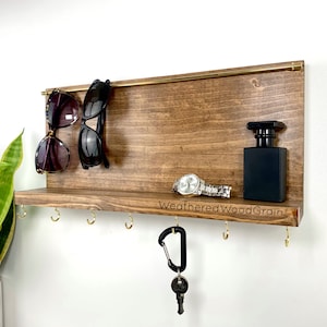 May include: A wooden wall mounted key holder with a shelf, hooks, and a brass rod. The shelf has a watch, a bottle of perfume, and the text "WeatheredWoodGrain" engraved on it. Two pairs of sunglasses are hanging from the brass rod.