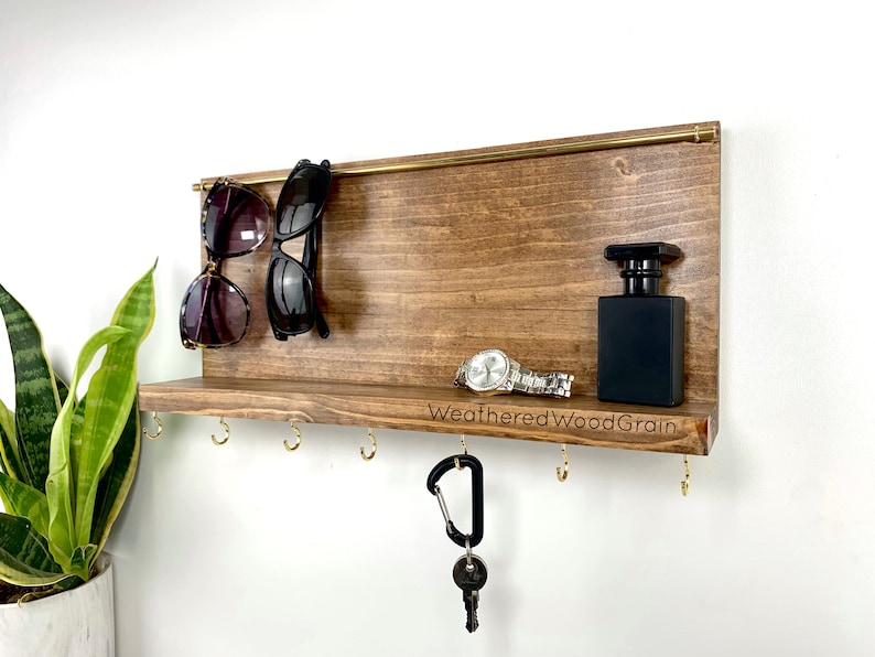 May include: A wooden wall mounted key holder with a shelf, hooks, and a brass rod. The shelf has a watch, a bottle of perfume, and the text "WeatheredWoodGrain" engraved on it. Two pairs of sunglasses are hanging from the brass rod.
