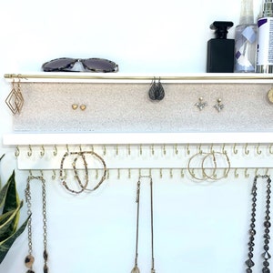 May include: White jewelry organizer with two shelves. The top shelf has a gold bar and a beige felt surface for storing earrings. The bottom shelf has a row of gold hooks for hanging necklaces and bracelets.