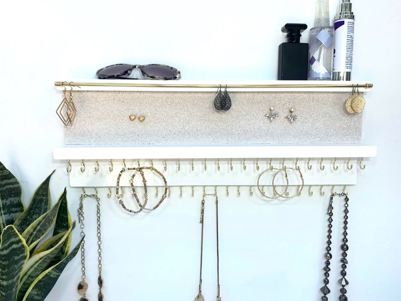 May include: White jewelry organizer with two shelves. The top shelf has a gold bar and a beige felt surface for storing earrings. The bottom shelf has a row of gold hooks for hanging necklaces and bracelets.