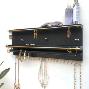 Puede incluir: Un organizador de joyas de pared en color negro con dos barras horizontales de metal dorado para pendientes, una barra negra inferior para pulseras y una fila de peque&ntilde;os ganchos dorados en el borde inferior para colgar collares y joyas.