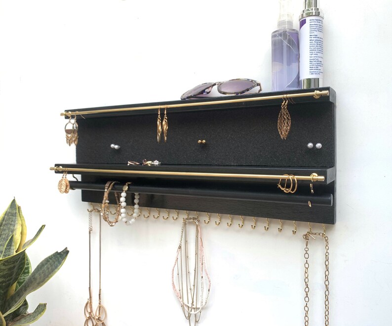 Puede incluir: Un organizador de joyas de pared en color negro con dos barras horizontales de metal dorado para pendientes, una barra negra inferior para pulseras y una fila de peque&ntilde;os ganchos dorados en el borde inferior para colgar collares y joyas.