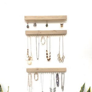 K&ouml;nnte beinhalten: Ein h&ouml;lzerner Schmuck-Organizer mit drei horizontalen Stangen, jede mit kleinen Haken. Verschiedene Halsketten und Ohrringe h&auml;ngen an den Haken. Das Holz hat eine helle, nat&uuml;rliche Oberfl&auml;che. Der Hintergrund ist eine schlichte wei&szlig;e Wand.