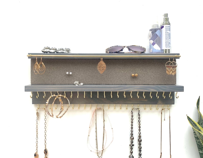 May include: A gray wooden jewelry organizer with gold hooks and a gray felt-lined top shelf. The organizer has a row of hooks for necklaces and a top shelf for earrings and other small jewelry.