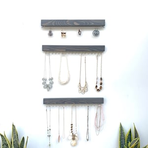 K&ouml;nnte beinhalten: Ein an der Wand befestigter Schmuck-Organizer mit drei grauen Holzleisten. Die obere Leiste h&auml;lt Ohrringe, w&auml;hrend die unteren Leisten Halsketten in verschiedenen Stilen und L&auml;ngen pr&auml;sentieren. Der Schmuck wird vor einer wei&szlig;en Wand ausgestellt.