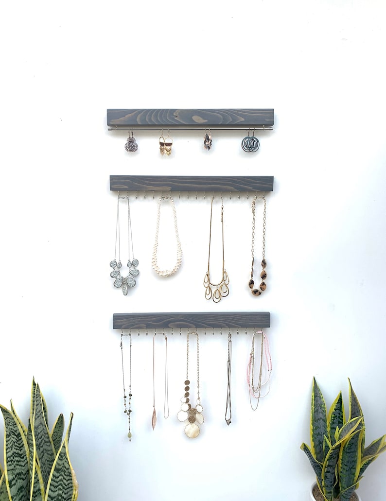 K&ouml;nnte beinhalten: Ein an der Wand befestigter Schmuck-Organizer mit drei grauen Holzleisten. Die obere Leiste h&auml;lt Ohrringe, w&auml;hrend die unteren Leisten Halsketten in verschiedenen Stilen und L&auml;ngen pr&auml;sentieren. Der Schmuck wird vor einer wei&szlig;en Wand ausgestellt.