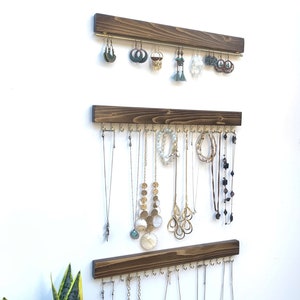 K&ouml;nnte beinhalten: Drei Schmuck-Organizer aus Holz an einer wei&szlig;en Wand. Der obere Organizer enth&auml;lt Ohrringe, w&auml;hrend die unteren beiden Halsketten pr&auml;sentieren. Das Holz ist dunkelbraun gebeizt und die Haken sind goldfarben.