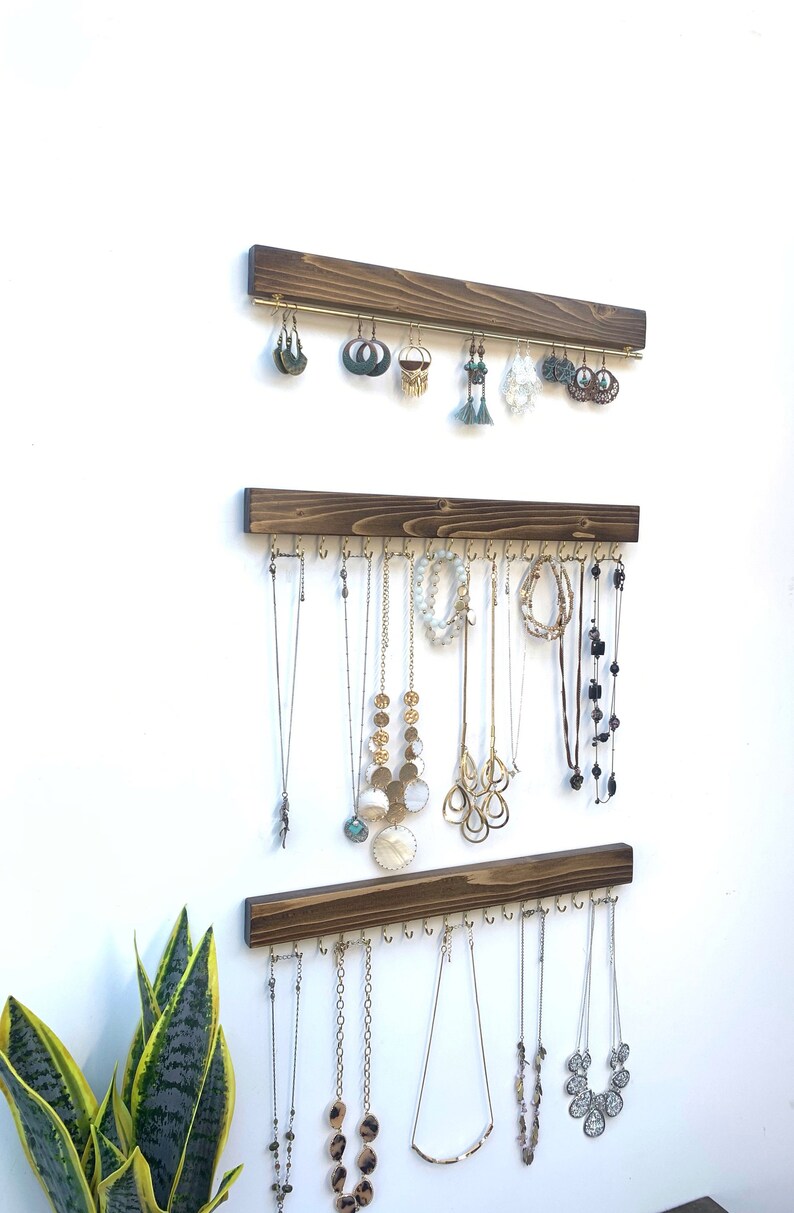 K&ouml;nnte beinhalten: Drei Schmuck-Organizer aus Holz an einer wei&szlig;en Wand. Der obere Organizer enth&auml;lt Ohrringe, w&auml;hrend die unteren beiden Halsketten pr&auml;sentieren. Das Holz ist dunkelbraun gebeizt und die Haken sind goldfarben.