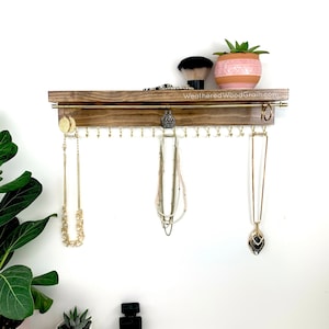 May include: A wooden jewelry organizer with a shelf and hooks for hanging necklaces and earrings. The organizer is stained a dark brown color and has a gold metal rod for hanging necklaces. There is a small potted succulent plant on the shelf.