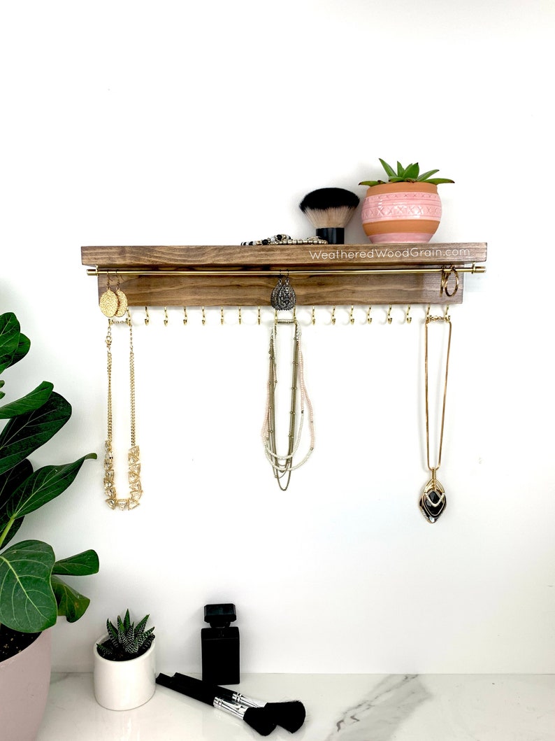 May include: A wooden jewelry organizer with a shelf and hooks for hanging necklaces and earrings. The organizer is stained a dark brown color and has a gold metal rod for hanging necklaces. There is a small potted succulent plant on the shelf.
