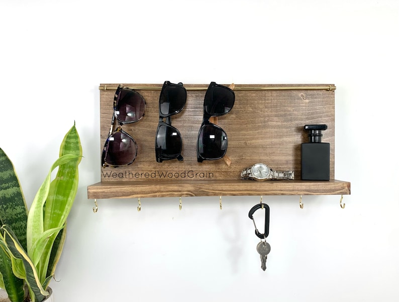 May include: A wooden wall-mounted organiser with hooks for keys and a shelf for sunglasses, a watch, and a small bottle. The shelf is stained a dark brown and has the text "WeatheredWoodGrain" printed on it.
