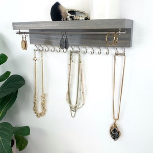 May include: A gray wooden jewelry organizer with a metal bar for hanging earrings and hooks for necklaces. The organizer is mounted on a white wall. The text "WeatheredWoodGrain.com" is printed on the organizer.