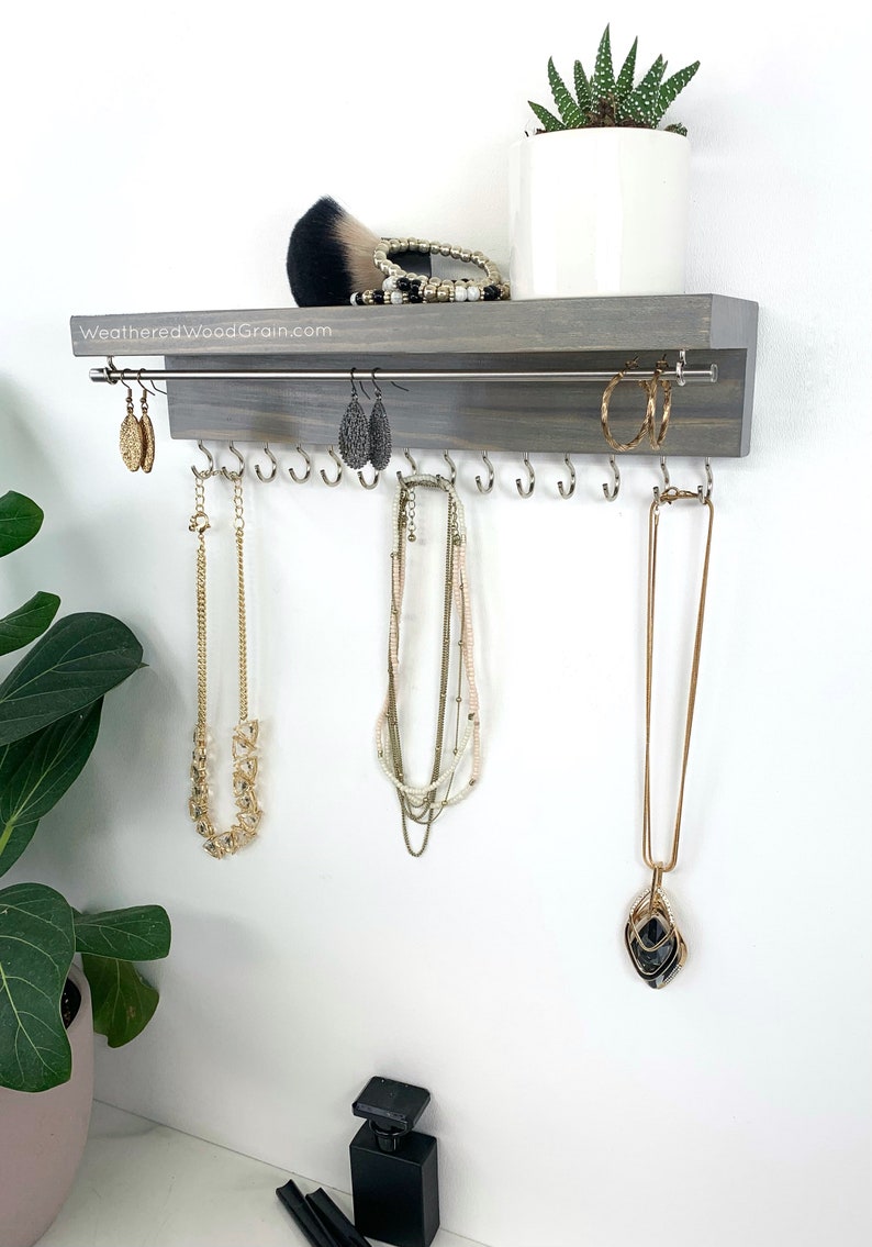 May include: A gray wooden jewelry organizer with a metal bar for hanging earrings and hooks for necklaces. The organizer is mounted on a white wall. The text "WeatheredWoodGrain.com" is printed on the organizer.