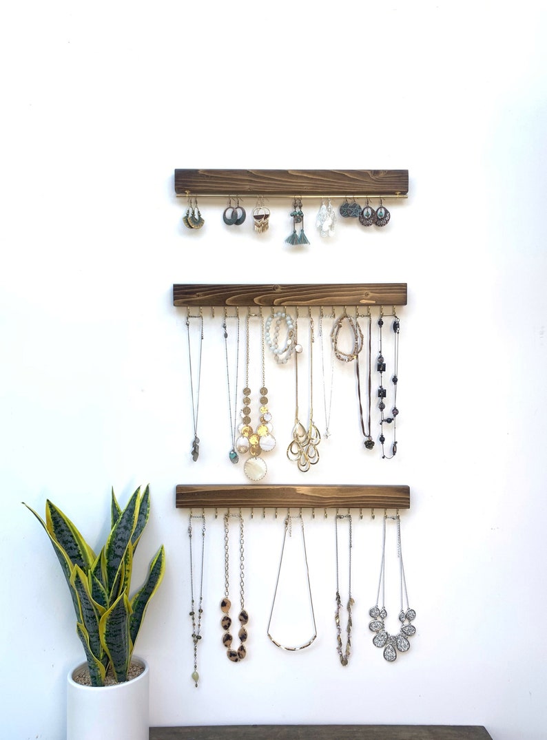 K&ouml;nnte beinhalten: Drei dunkle, braune Holzschmuck-Organizer an einer wei&szlig;en Wand, die Halsketten und Ohrringe zeigen. Jeder Organizer hat mehrere Haken. Eine Topf-Schlangenpflanze befindet sich in der linken unteren Ecke.