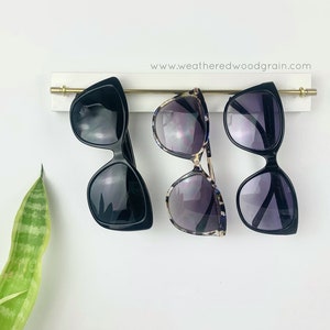 May include: A white wall-mounted rack with a horizontal gold metal rod holding three pairs of sunglasses. The rack features the text www.weatheredwoodgrain.com. Two pairs of sunglasses have solid black frames, and one pair has a tortoiseshell pattern.