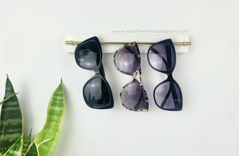 May include: A white wall-mounted rack with a horizontal gold metal rod holding three pairs of sunglasses. The rack features the text www.weatheredwoodgrain.com. Two pairs of sunglasses have solid black frames, and one pair has a tortoiseshell pattern.