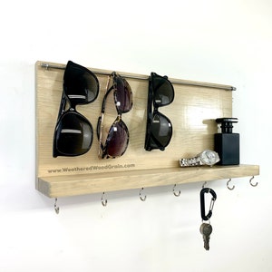 May include: A light wood wall mounted shelf with a metal rod for hanging sunglasses. The shelf has hooks for keys and a small shelf for other items. The text "www.WeatheredWoodGrain.com" is printed on the shelf.