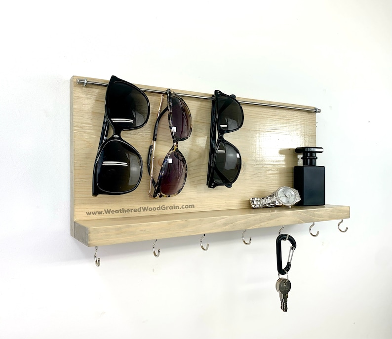 May include: A light wood wall mounted shelf with a metal rod for hanging sunglasses. The shelf has hooks for keys and a small shelf for other items. The text "www.WeatheredWoodGrain.com" is printed on the shelf.