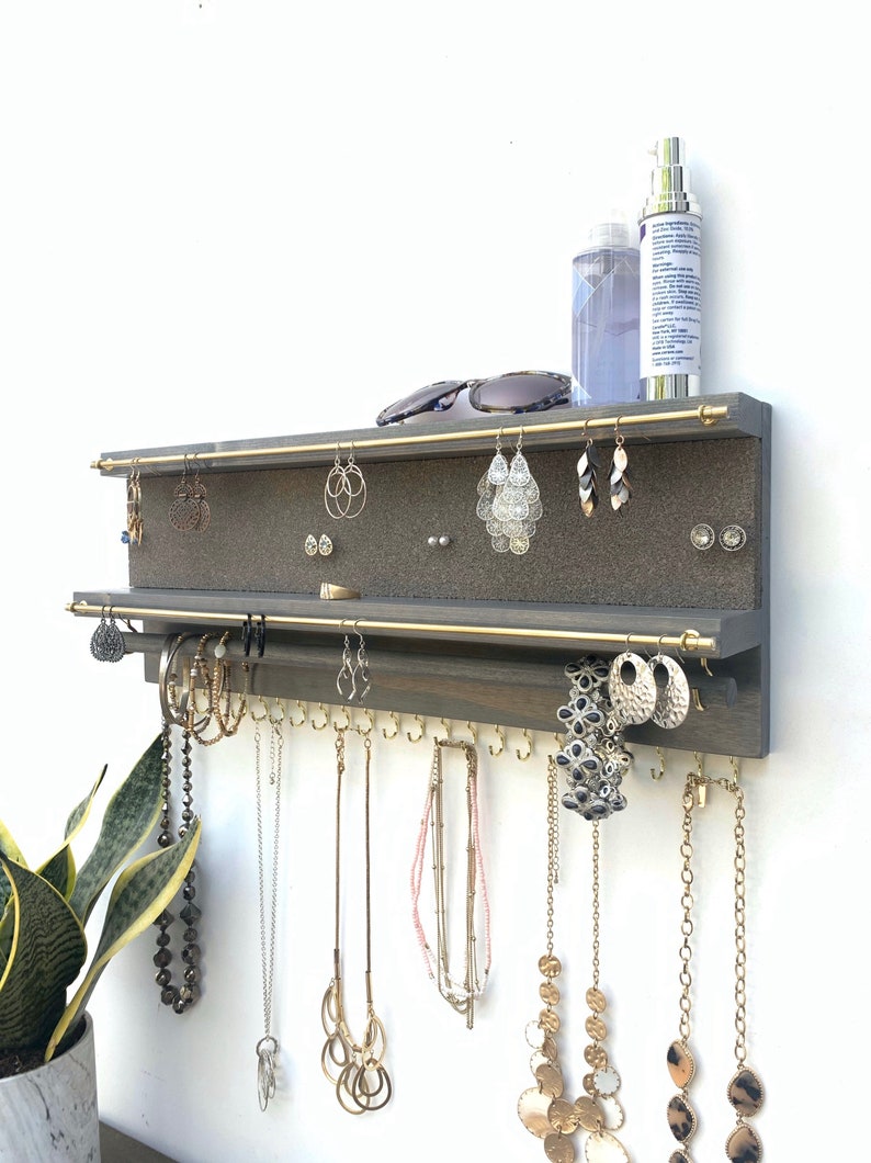 Puede incluir: Un organizador de joyas de madera gris montado en la pared con dos varillas horizontales de lat&oacute;n para pendientes, un panel de corcho para pendientes de bot&oacute;n y una fila de ganchos de lat&oacute;n en la parte inferior para colgar collares y pulseras.