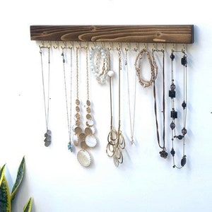 Pu&ograve; includere: Un organizzatore di gioielli da parete rettangolare in legno tinto marrone scuro, dotato di una fila di ganci in metallo dorato distanziati uniformemente. Diverse collane, bracciali e catenine di varie lunghezze e stili sono appesi ai ganci.