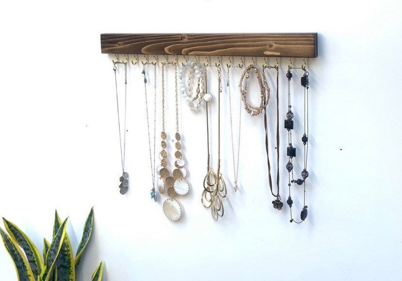 Pu&ograve; includere: Un organizzatore di gioielli da parete rettangolare in legno tinto marrone scuro, dotato di una fila di ganci in metallo dorato distanziati uniformemente. Diverse collane, bracciali e catenine di varie lunghezze e stili sono appesi ai ganci.