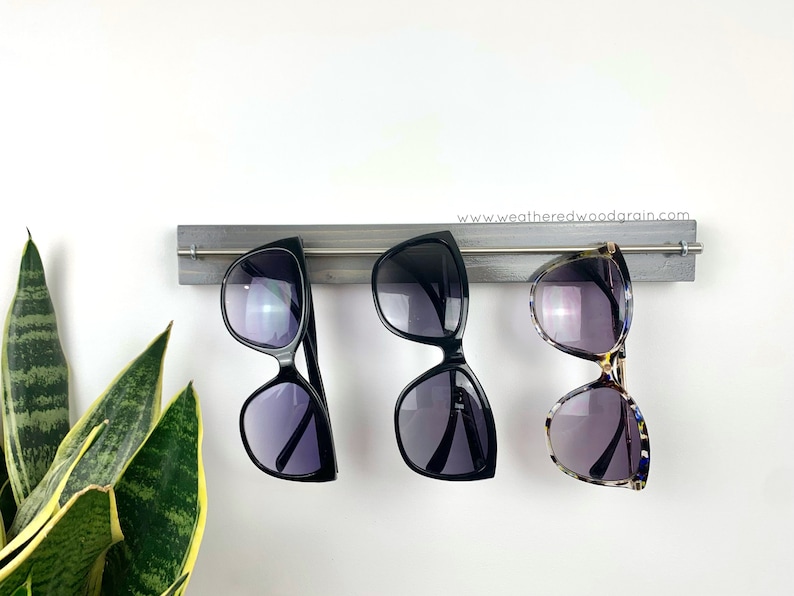 May include: A grey wooden wall-mounted rack featuring a horizontal silver metal rod. Three pairs of sunglasses are hanging from the rod: one black pair on the left, one black pair in the centre, and one tortoiseshell pair on the right. The text www.weatheredwoodgrain.com is printed on the wood.