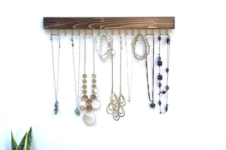 Pu&ograve; includere: Un organizer da parete per gioielli in legno tinto marrone scuro, caratterizzato da una tavola orizzontale con molteplici ganci in metallo dorato. Diverse collane, bracciali e catenine di varie lunghezze e stili sono appesi ai ganci.