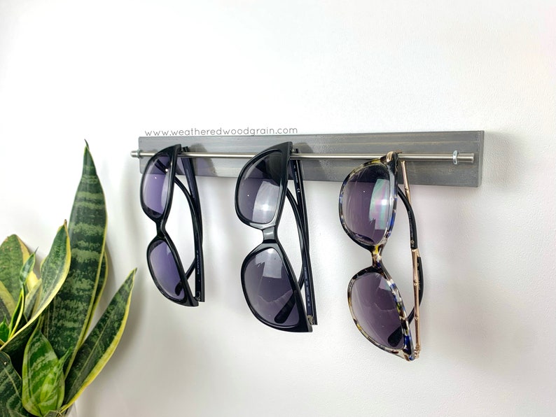 May include: A grey wooden wall-mounted rack featuring a horizontal silver metal rod for hanging eyewear. Three pairs of sunglasses are suspended from the rod. The text www.weatheredwoodgrain.com is printed on the top edge of the wooden board.