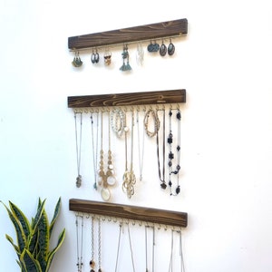 K&ouml;nnte beinhalten: Drei h&ouml;lzerne Schmuckorganisatoren an einer wei&szlig;en Wand, jeder mit einer Reihe von Haken, die verschiedene Halsketten und Ohrringe halten. Das Holz ist dunkelbraun gebeizt, und der Schmuck umfasst silberne, goldene und Perlenst&uuml;cke.