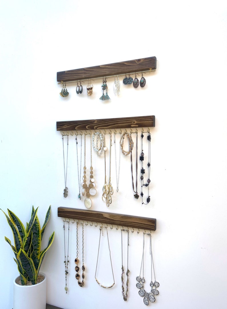 K&ouml;nnte beinhalten: Drei h&ouml;lzerne Schmuckorganisatoren an einer wei&szlig;en Wand, jeder mit einer Reihe von Haken, die verschiedene Halsketten und Ohrringe halten. Das Holz ist dunkelbraun gebeizt, und der Schmuck umfasst silberne, goldene und Perlenst&uuml;cke.