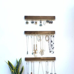 K&ouml;nnte beinhalten: Drei h&ouml;lzerne Schmuck-Organizer an einer wei&szlig;en Wand, die jeweils verschiedene Ohrringe und Halsketten halten. Die Organizer sind dunkelbraun gebeizt und haben kleine Haken zum Aufh&auml;ngen von Schmuck. Eine Topf-Bogenhanf befindet sich in der linken unteren Ecke.