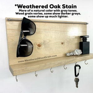 May include: A weathered oak wood key holder with a shelf and hooks. The shelf has a watch, a bottle of perfume, and sunglasses on it. The text "*Weathered Oak Stain" is at the top of the key holder. The text "More of a natural colour with grey tones. Wood grain varies, some show darker greys, some show up much lighter." is below the title. The text "www.WeatheredWoodGrain.com" is at the bottom of the key holder.