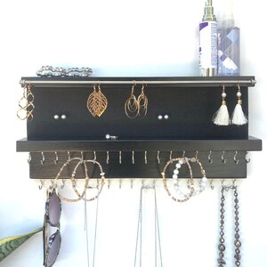 May include: Black wooden jewelry organizer with hooks for necklaces and earrings. The organizer has a top shelf for storing bracelets and other small items.