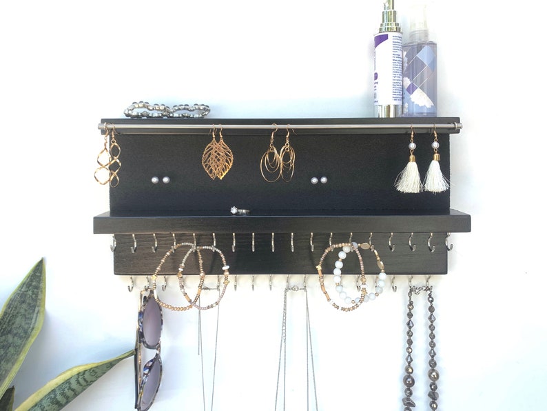 May include: Black wooden jewelry organizer with hooks for necklaces and earrings. The organizer has a top shelf for storing bracelets and other small items.