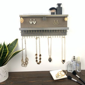 May include: Gray wooden jewelry organizer with a top shelf for earrings and a bottom shelf with hooks for necklaces. The organizer is mounted on a white wall. The top shelf has a brown felt lining. The bottom shelf has gold hooks. There are several necklaces and earrings hanging on the organizer. The text "www.WeatheredWoodGrain.com" is printed on the top shelf.