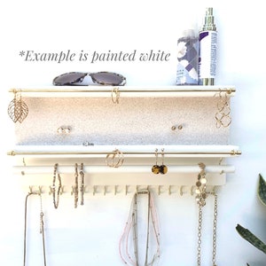Puede incluir: Un organizador de joyas de pared blanco con dos barras horizontales de metal dorado para pendientes, una barra blanca inferior con ganchos para collares y pulseras, y una superficie de estante texturizada. El texto *Example is painted white* es visible.