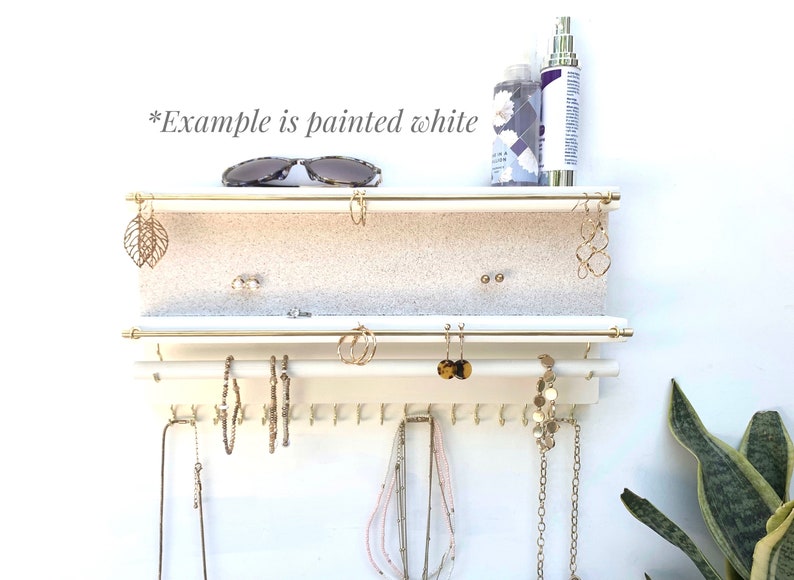 Puede incluir: Un organizador de joyas de pared blanco con dos barras horizontales de metal dorado para pendientes, una barra blanca inferior con ganchos para collares y pulseras, y una superficie de estante texturizada. El texto *Example is painted white* es visible.