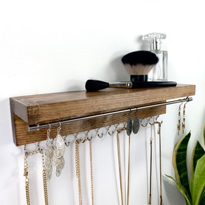 May include: A wooden shelf with a metal rod for hanging jewelry. The shelf has a dark brown stain and is mounted on a white wall. There are several necklaces and earrings hanging from the rod. A makeup brush and a bottle of perfume are on the shelf.