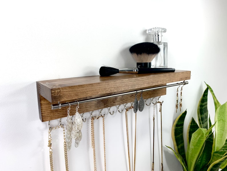 May include: A wooden shelf with a metal rod for hanging jewelry. The shelf has a dark brown stain and is mounted on a white wall. There are several necklaces and earrings hanging from the rod. A makeup brush and a bottle of perfume are on the shelf.