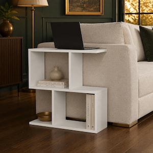 May include: A white S-shaped side table with multiple open storage compartments. The top surface holds a black laptop, while the middle and bottom shelves contain a small beige vase, a gold bowl, and several books titled Live Beautifully and The Kinfolk.