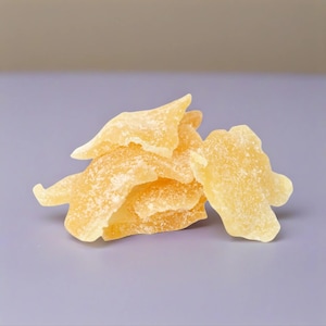 May include: A small pile of translucent, golden-yellow crystallized ginger slices. Each piece has an irregular, jagged shape with a visible coating of fine, white sugar granules on the surface, giving the edges a textured and slightly sparkling appearance.