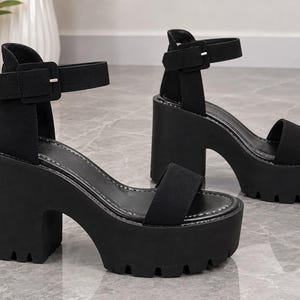 Op de afbeelding: Een paar zwarte plateausandalen met een dikke blokhak, een grove profielzool en een enkelbandje met een vierkante gesp. De sandalen hebben een brede band over de tenen en zijn afgewerkt met een glad, mat materiaal aan de buitenzijde.