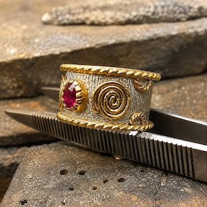 Pode incluir: Um anel de banda larga com um centro prateado texturizado, bordas douradas estilo corda e detalhes dourados em espiral. Uma única gema redonda, facetada e de cor vermelho-rosada intensa está engastada num bisel dourado na frente do anel.