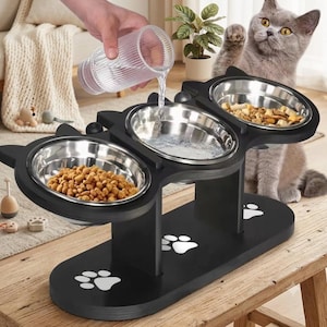 May include: A black elevated pet feeder stand with three stainless steel bowls, featuring cat-ear shaped sides and white paw print designs on the base. Two bowls are filled with dry kibble, while the center bowl is being filled with water.