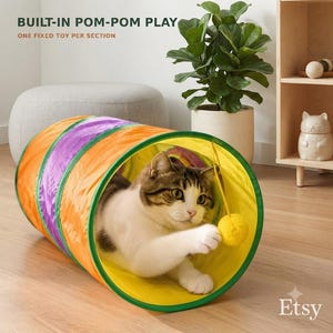 May include: A collapsible cat tunnel featuring orange, purple, and yellow fabric sections with green trim. A brown and white tabby cat is inside the tunnel, batting at a yellow pom-pom toy attached to the interior. Text reads BUILT-IN POM-POM PLAY.