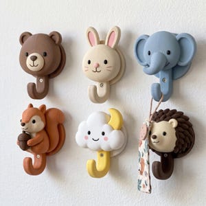 May include: A set of six wall-mounted hooks featuring animal and nature designs, including a brown bear, a cream rabbit, a blue elephant, an orange squirrel holding an acorn, a white cloud with a yellow moon, and a brown hedgehog with a hanging tag.