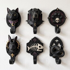 Puede incluir: Un conjunto de seis ganchos de pared negros con motivos animales esculpidos, que incluyen un gato con collar de media luna, un perro con alas de murciélago, una huella con una calavera, un conejo, un esqueleto de pez y una serpiente enroscada.