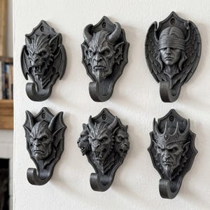 May include: A set of six dark gray wall-mounted hooks, each featuring a unique sculpted head design including a dragon, a horned demon, a blindfolded figure with wings, a gargoyle, a three-headed creature, and a crowned figure with a goatee.