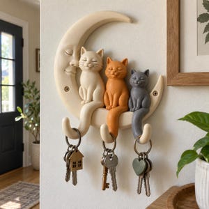 May include: A wall-mounted key holder shaped like a crescent moon with a face, featuring three sculpted cats in cream, orange, and grey sitting on the moon. Each cat has a tail that forms a hook, holding metal key rings with various keys attached.