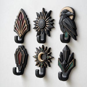 Peut inclure: Un ensemble de six patères décoratives aux motifs géométriques noirs, dorés et avec des accents rouge foncé ou vert. Les formes incluent un éventail, un soleil, un corbeau, un bouclier, un croissant de lune et un groupe de cristaux.